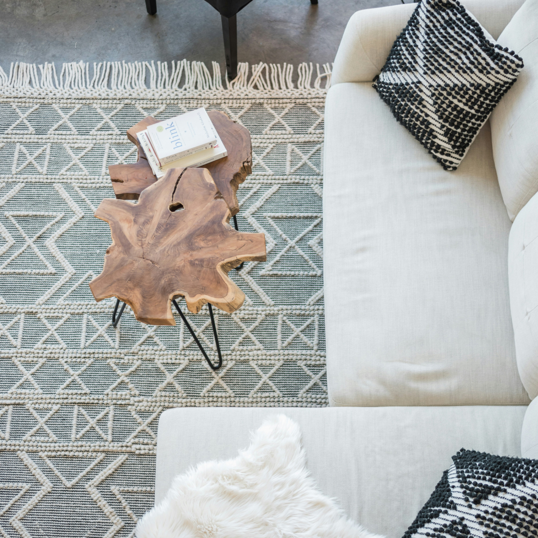 Overhead view of a cozy living room corner with a natural wood slab coffee table on hairpin legs, a beige sofa with patterned throw pillows, and a geometric patterned rug with a fringe border.