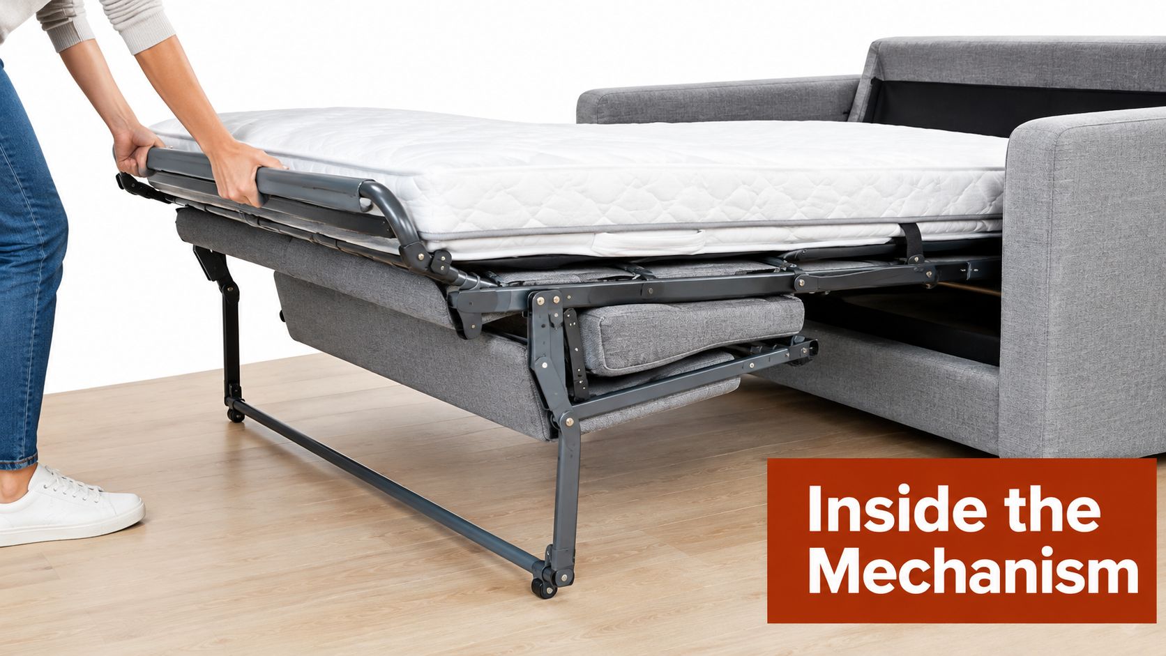 A person pulling open a gray sleeper sofa mechanism to reveal a folded mattress on a wooden floor.